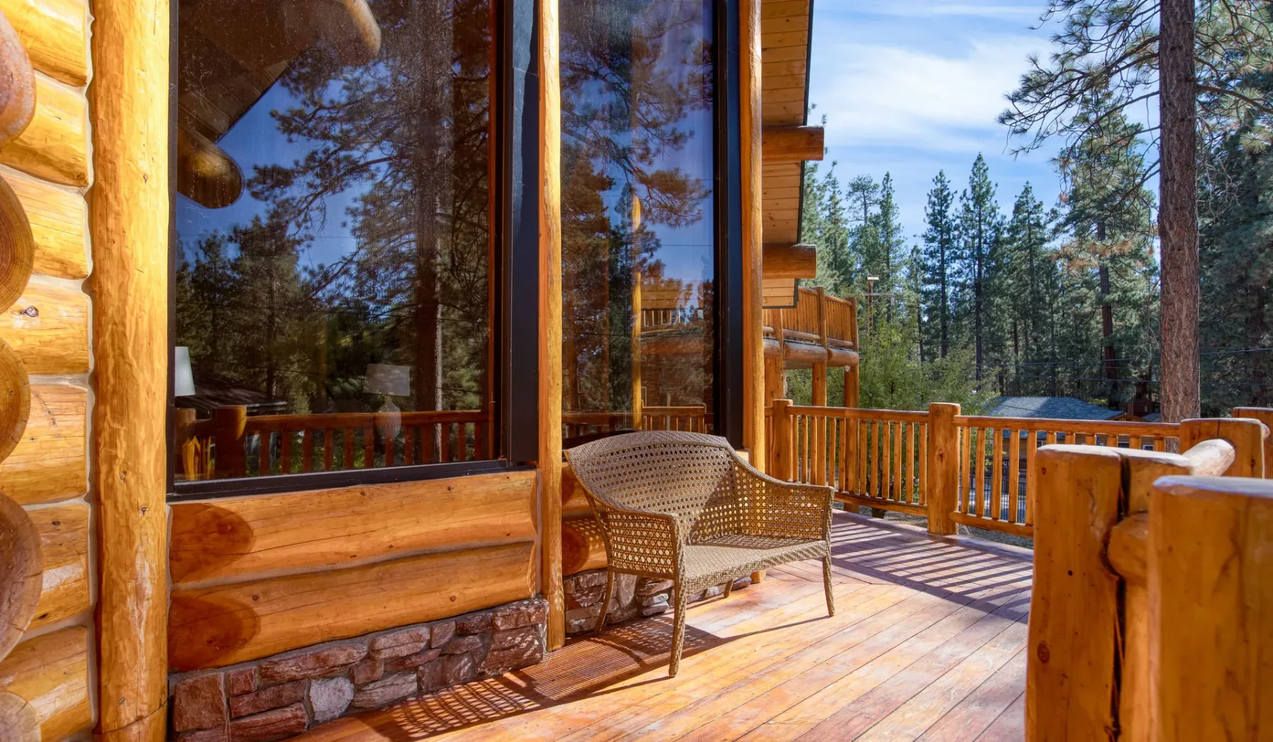 A wooden deck beside a cabin’s large windows overlooks a pine forest.
