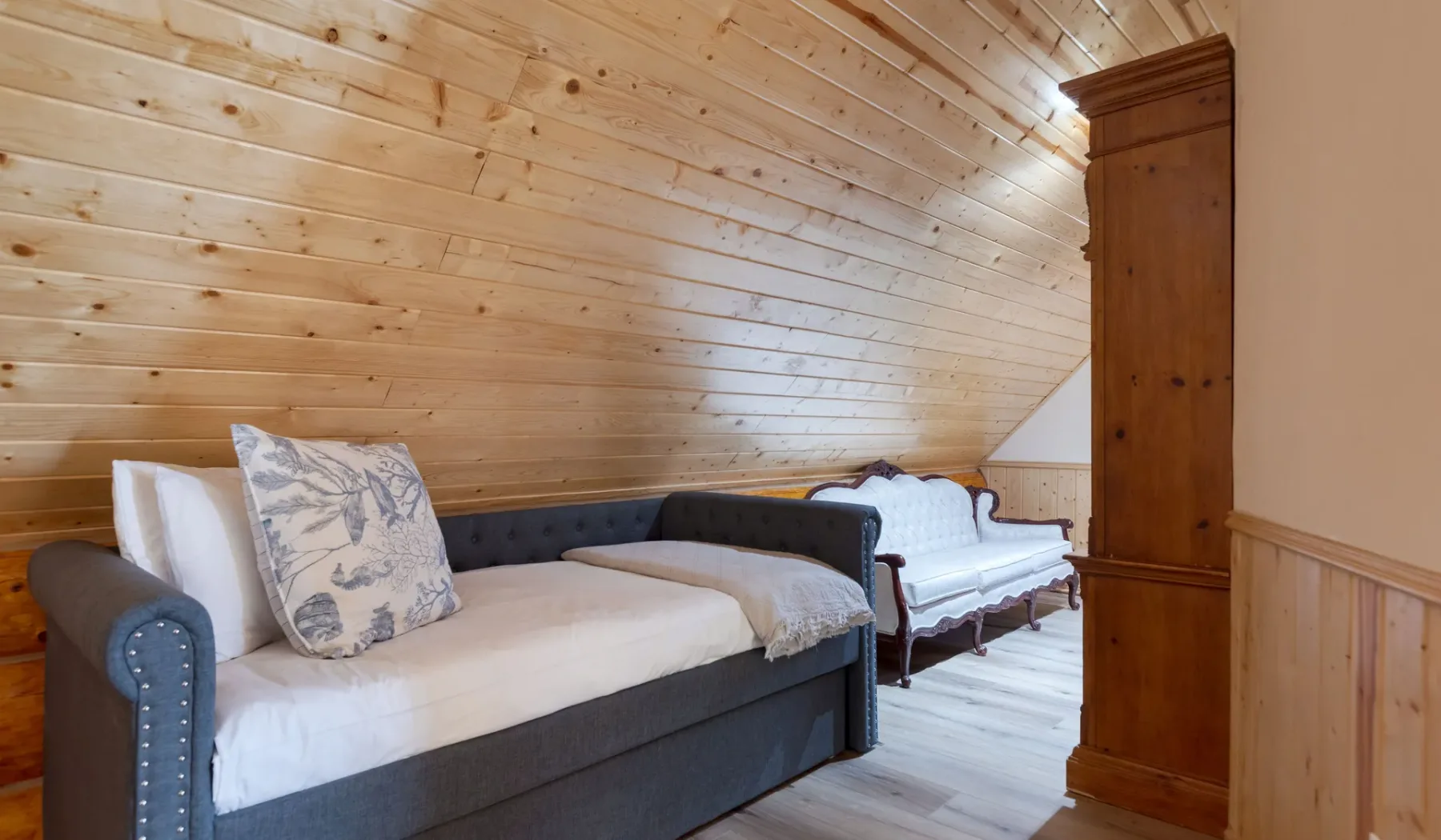 A sitting area with a dark upholstered daybed and a white settee beneath a sloped wood-paneled ceiling.