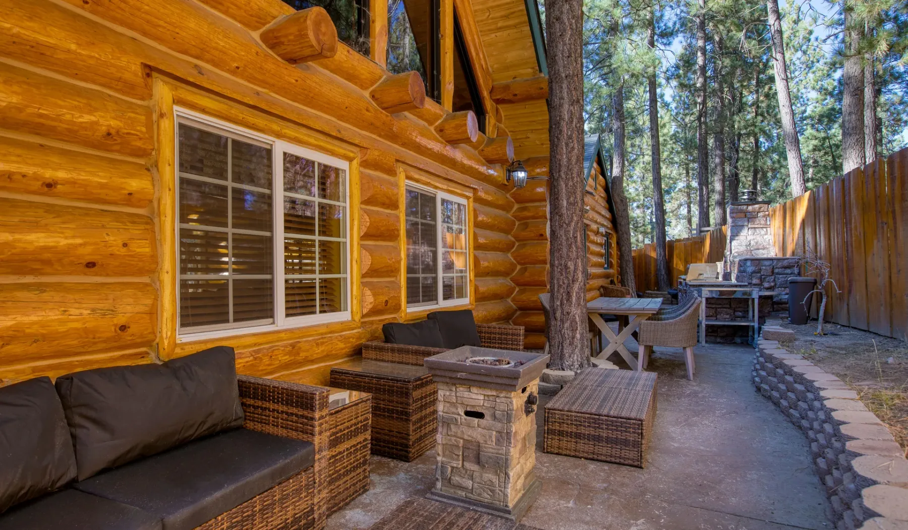 An outdoor patio beside a log cabin, with seating and a wooded view.
