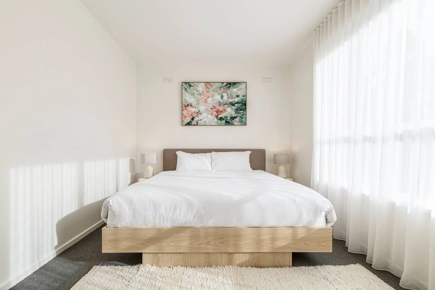 A bright bedroom features a neatly made bed beneath a floral painting, with sheer curtains along the window.