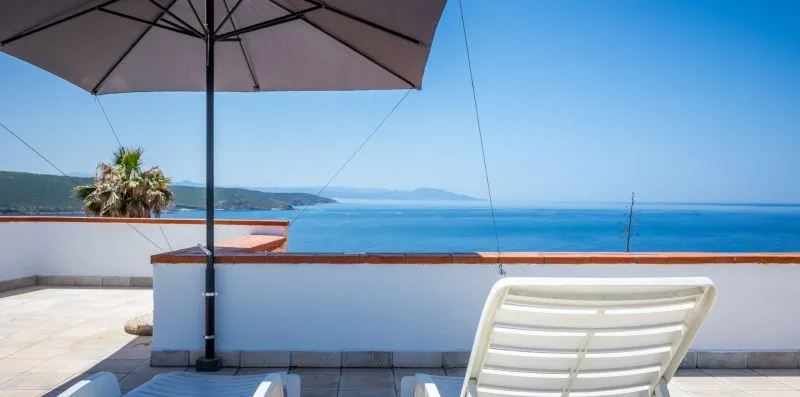 A seaside terrace overlooks the water, with a lounge chair beneath a large umbrella.
