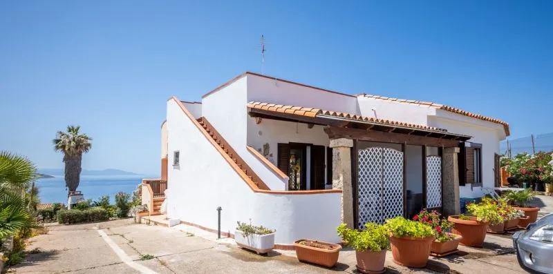 A white stucco house with a tiled roof, potted plants, and a view of the sea.