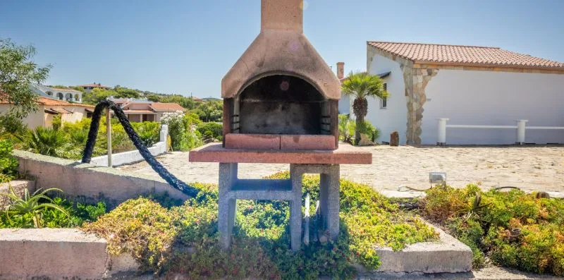 An outdoor barbecue grill stands beside a paved courtyard with garden beds.