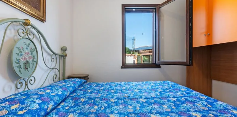 A bedroom with a bed covered in blue floral bedding beside an open window.