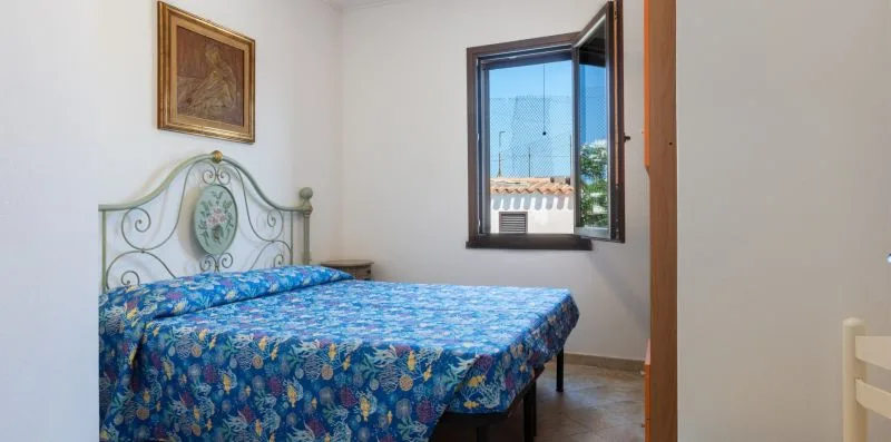 A bedroom with a bed covered in blue floral bedding beside an open window.
