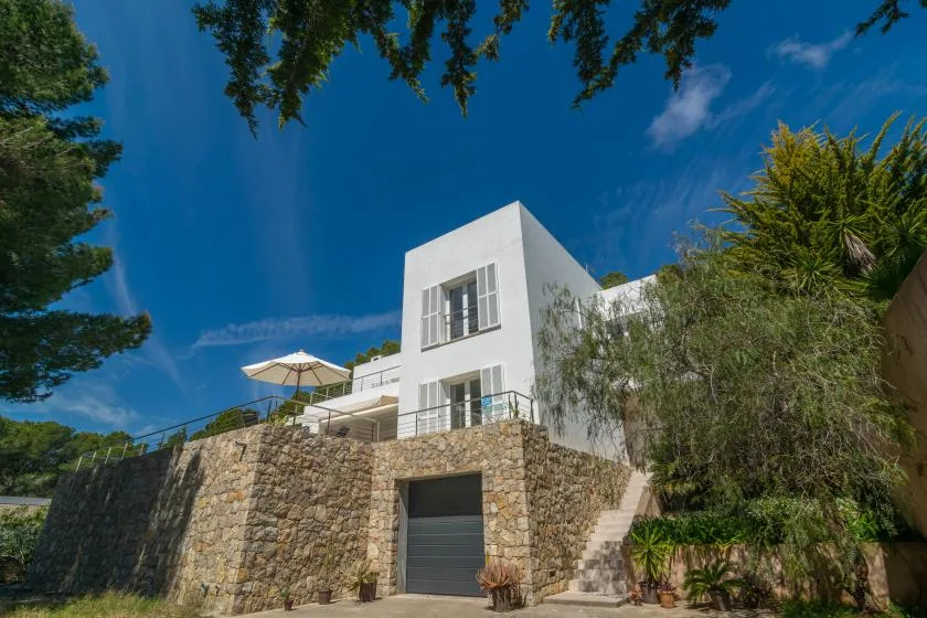 A white modern house rises above a stone retaining wall beneath a blue sky.