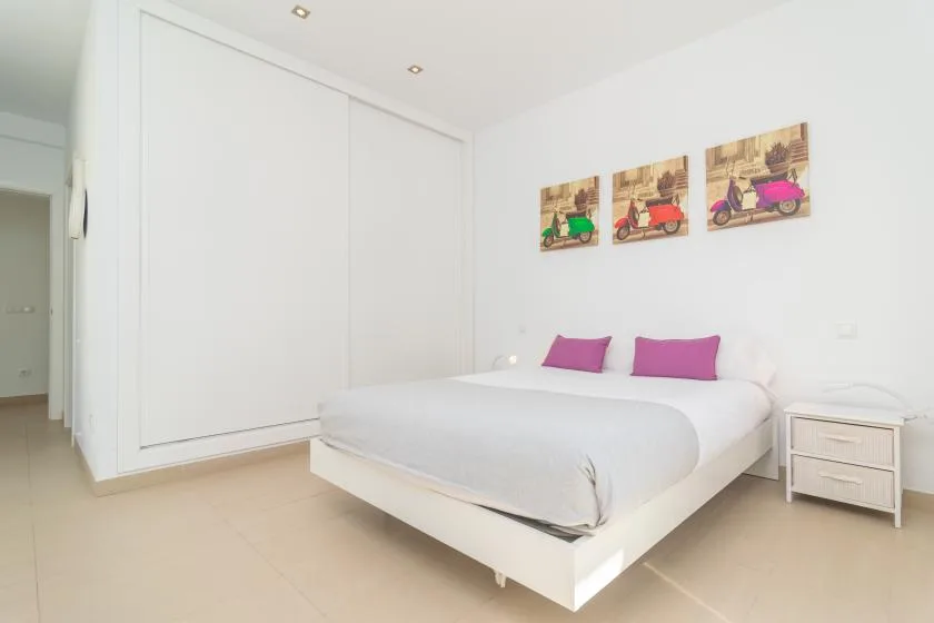 A bright bedroom with a white bed, purple pillows, and colorful wall art.