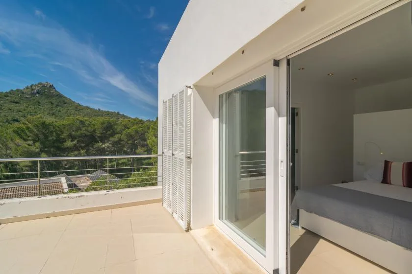 A sunny terrace opens onto a bedroom, with green hills visible beyond the railing.