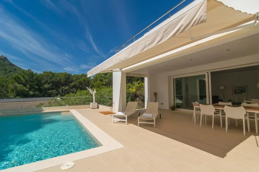A poolside patio with outdoor seating and a dining table beneath a broad awning.