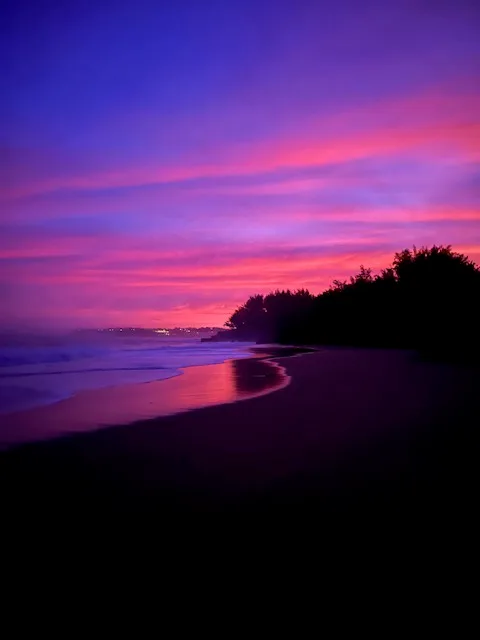 Una playa bordea el agua bajo un atardecer de tonos rosa y morado.
