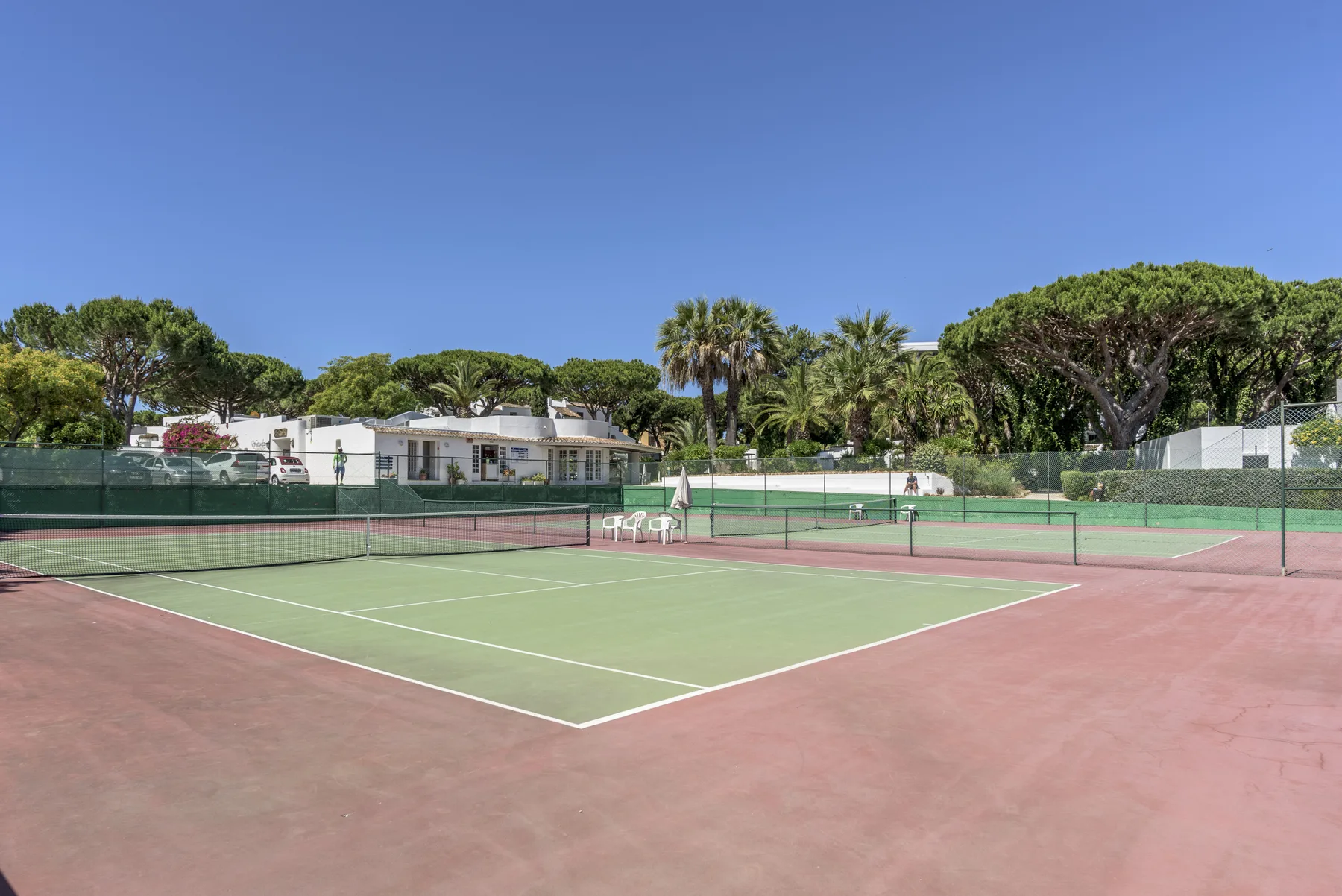 Outdoor tennis court with green playing surfaces and trees in the background.