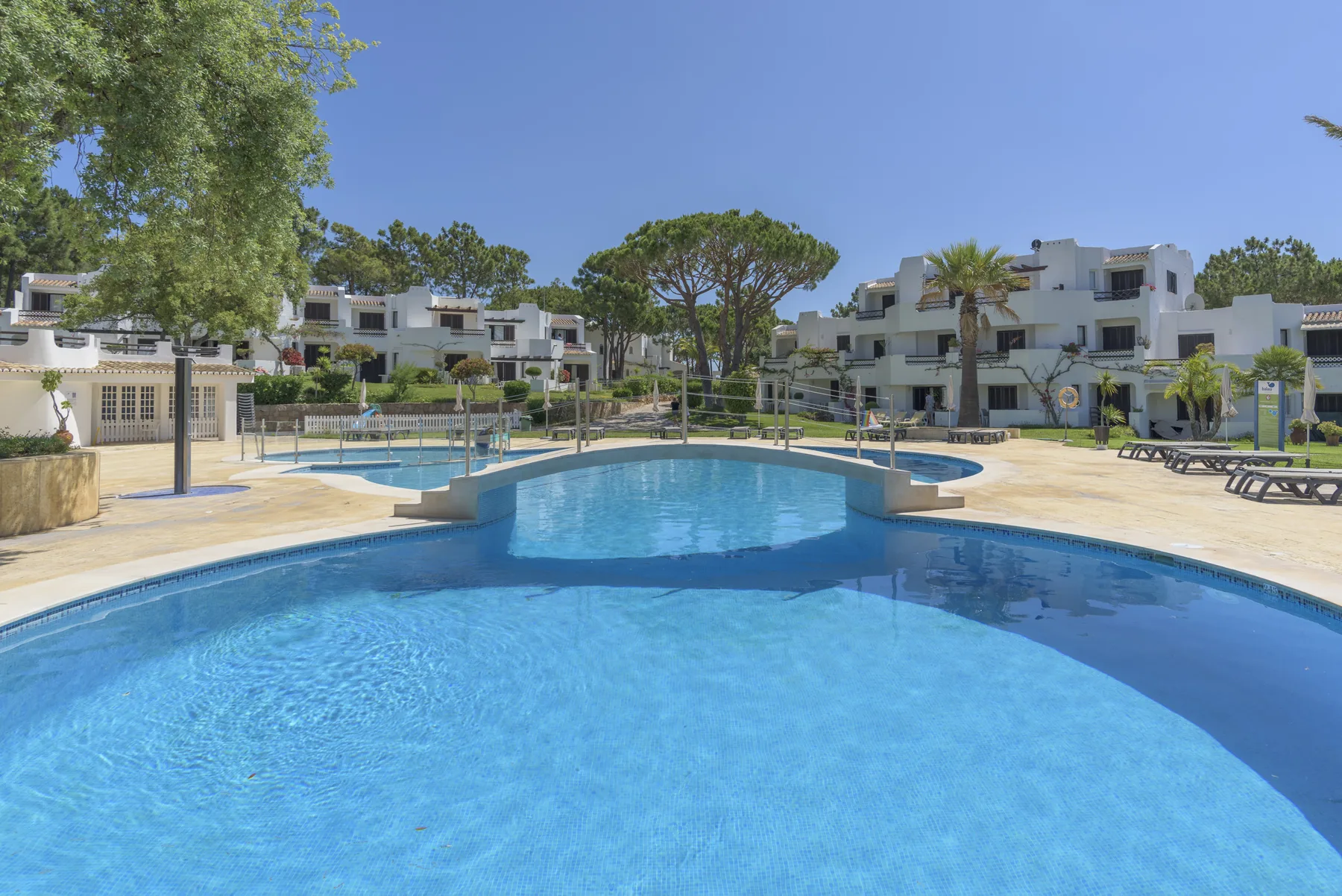 A swimming pool sits among low-rise buildings, trees, and paved poolside areas.