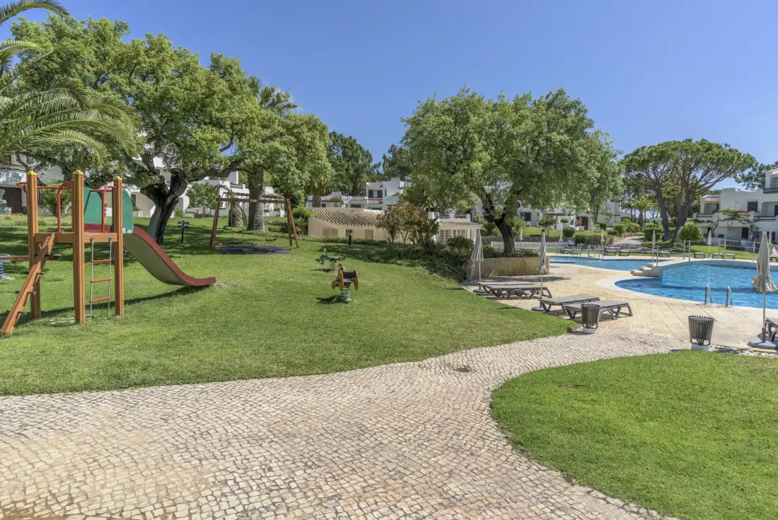 A landscaped outdoor recreation area with a playground, trees, and a swimming pool.