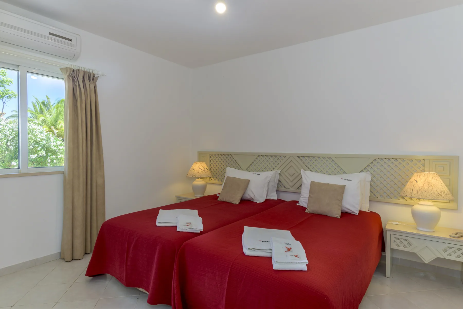 A bedroom with red bedspreads and a window overlooking greenery.