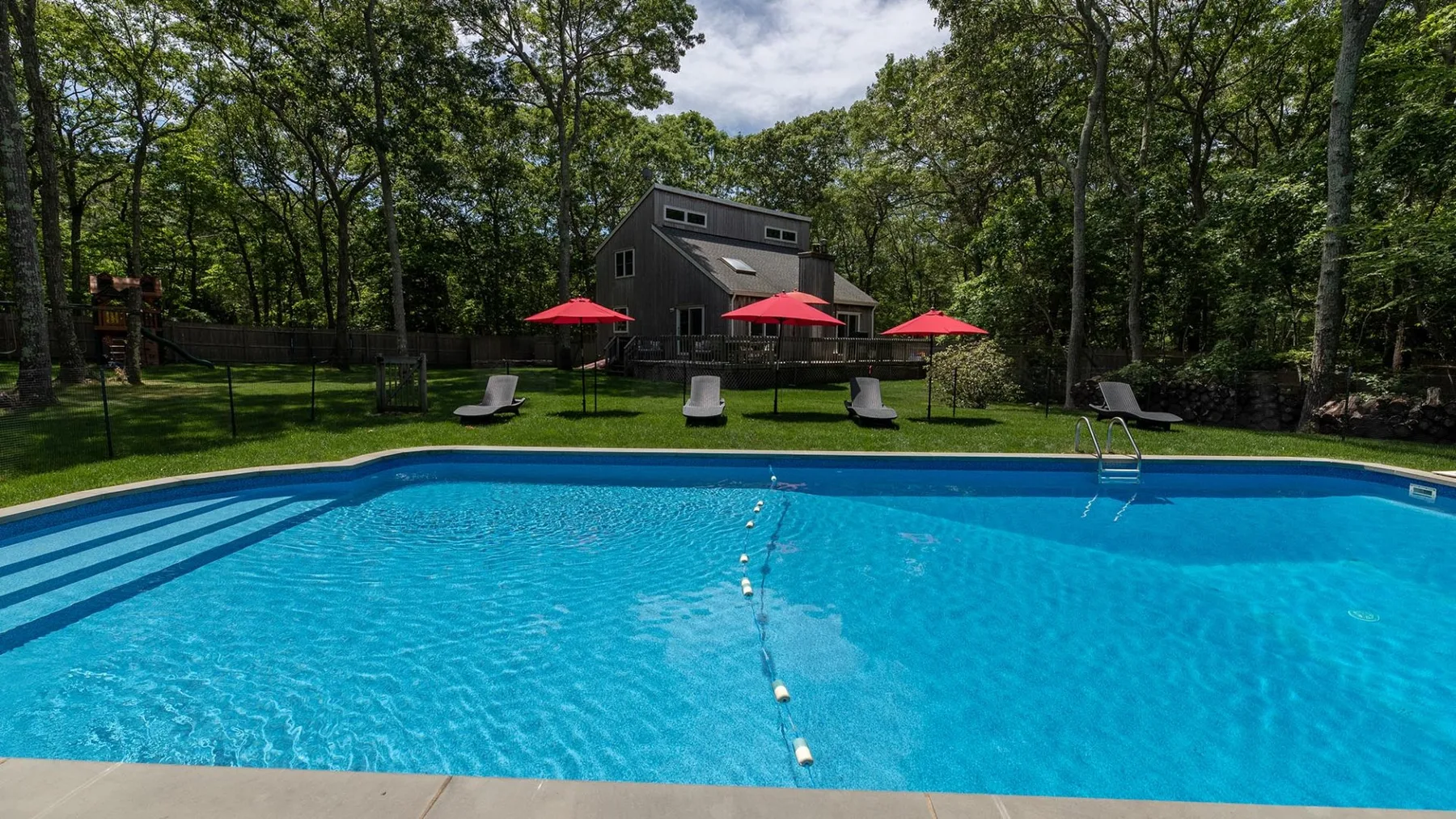 Una piscina frente a una casa oscura, rodeada de árboles y sombrillas rojas.