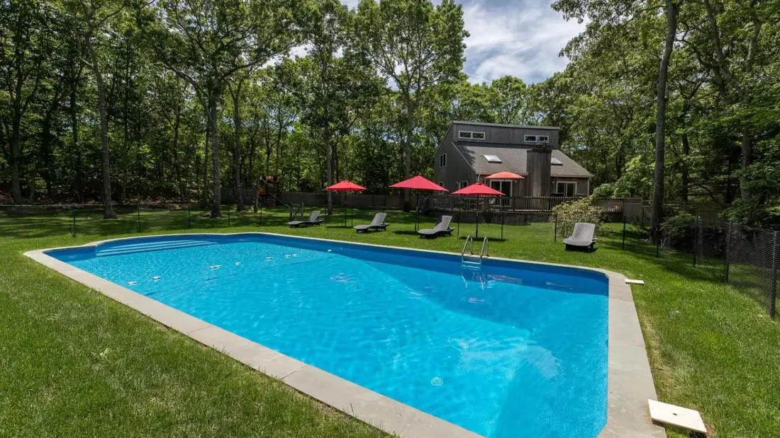 Una piscina está en el césped junto a una casa rodeada de árboles.