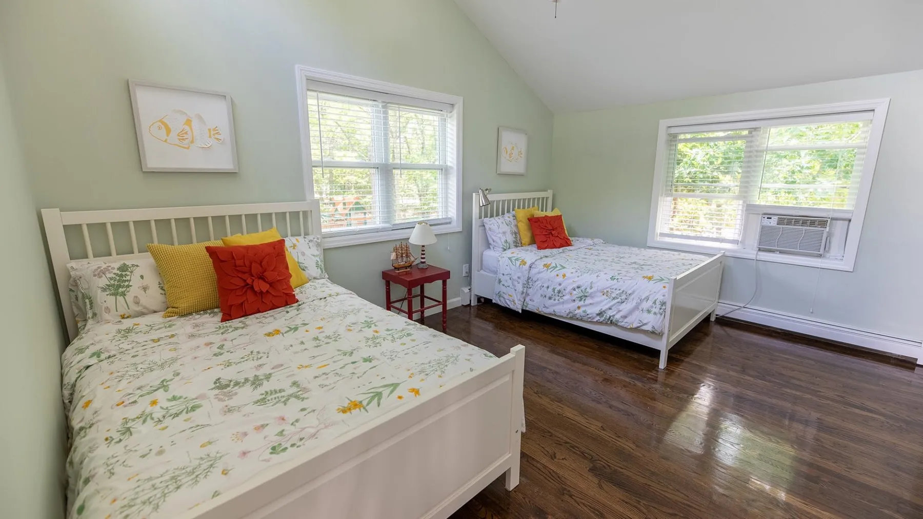 Un dormitorio luminoso con dos camas, ropa de cama floral y ventanas con vistas a la vegetación.