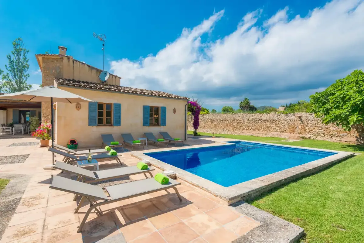 A sunny poolside patio beside a house, with lounge seating and a green lawn.