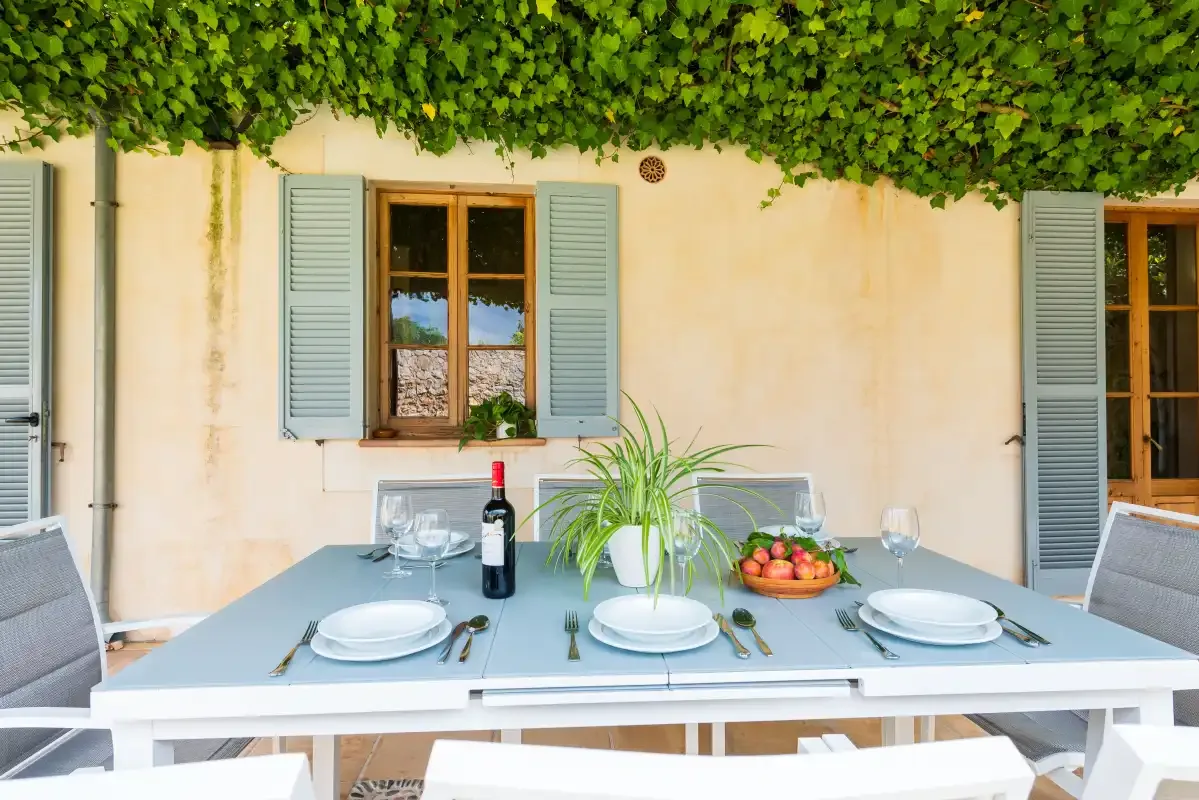 An outdoor dining table sits beneath leafy vines beside shuttered windows.