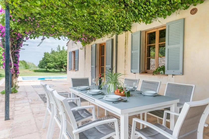 An outdoor dining area sits beneath leafy vines beside a house.