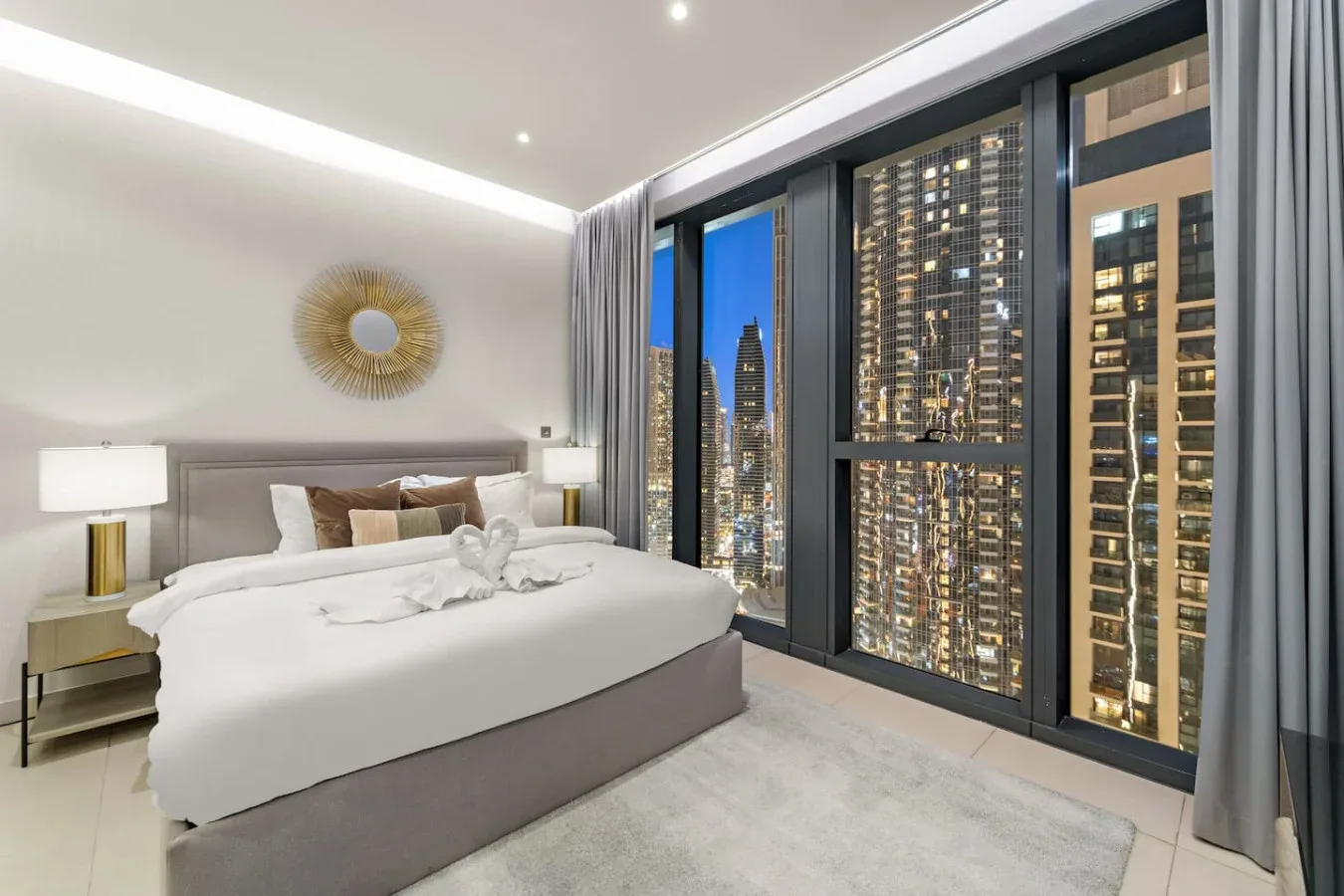 A modern bedroom with a neatly made bed beside floor-to-ceiling windows overlooking city buildings.