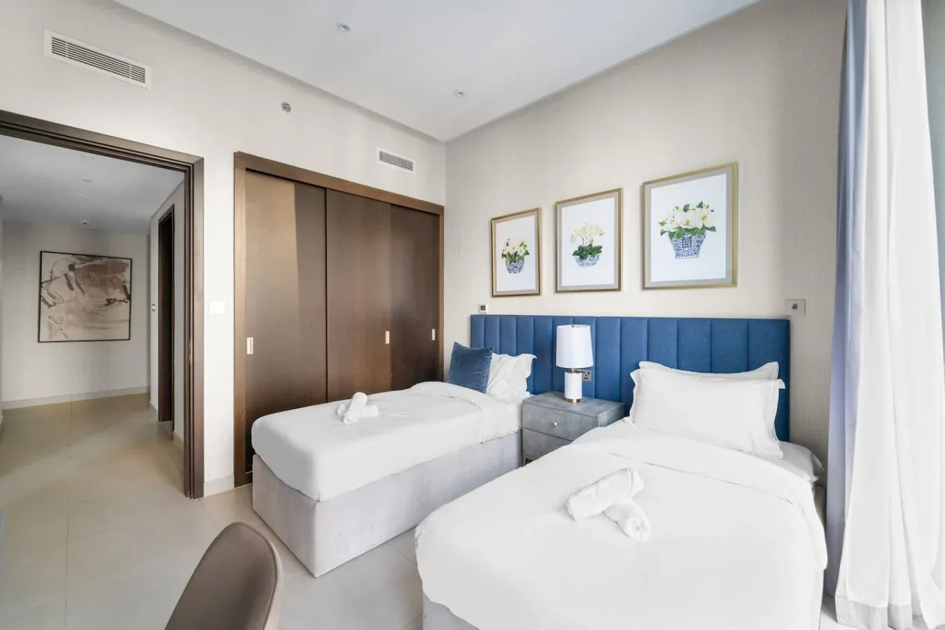 A bedroom with two beds, a blue headboard, and framed floral prints.