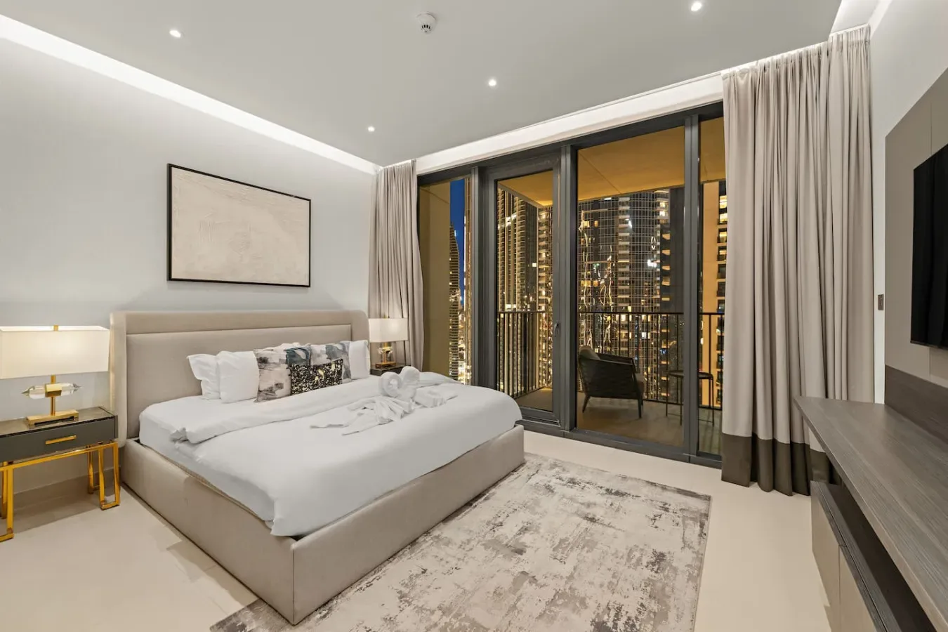 A modern bedroom with a neatly made bed and floor-to-ceiling windows overlooking city lights.