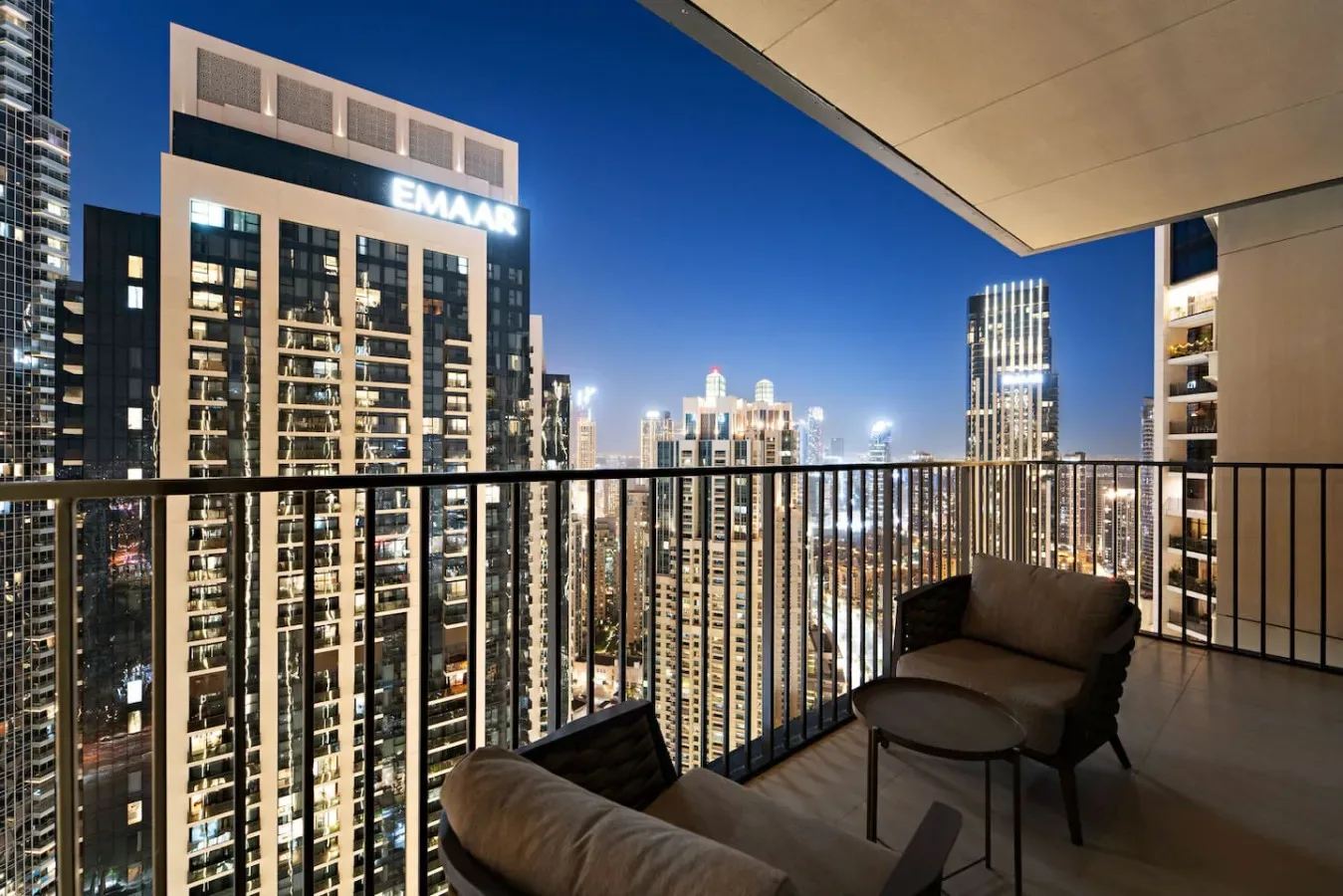 A balcony overlooks a brightly lit city skyline at night, with outdoor seating.