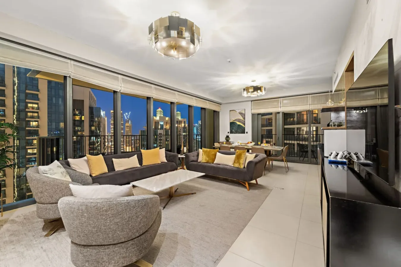 A modern living room with seating, a dining area, and large windows overlooking city buildings.