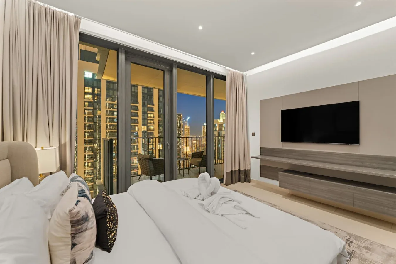 A bedroom with a large bed facing a wall-mounted television and city views.
