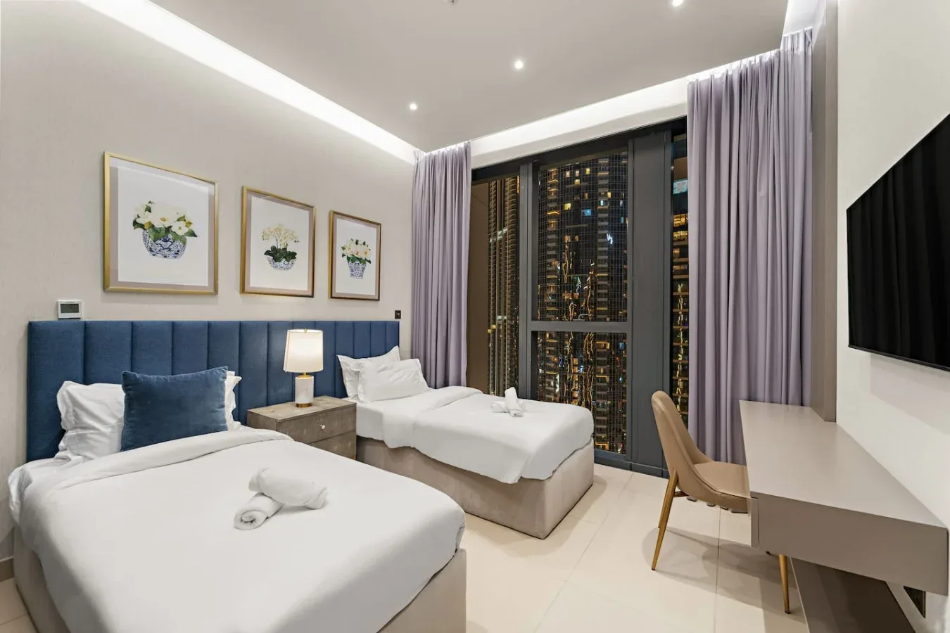 A bedroom with two beds, framed artwork, and a desk beside the window.