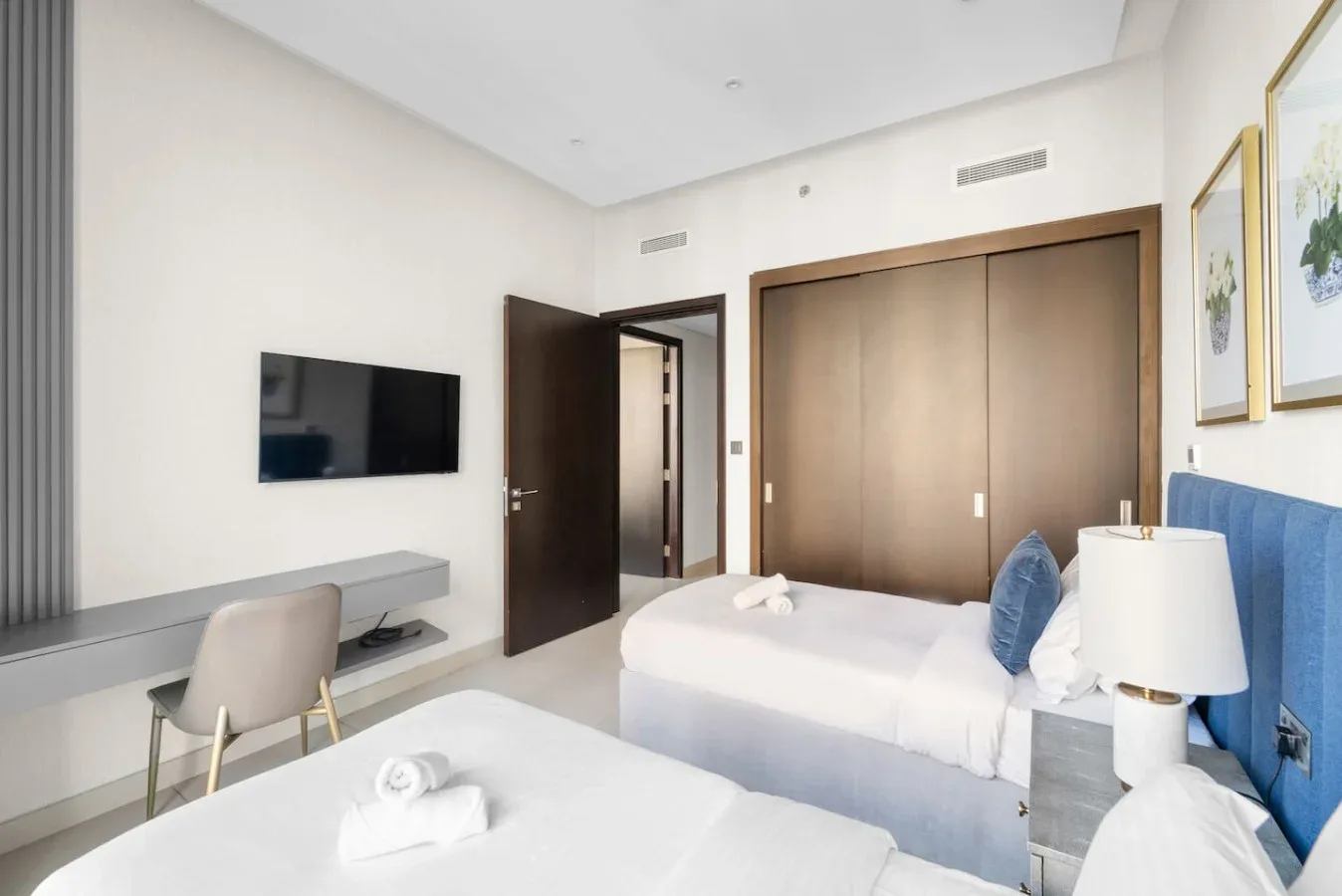 A bright bedroom has two beds, a desk, and a wall-mounted television.