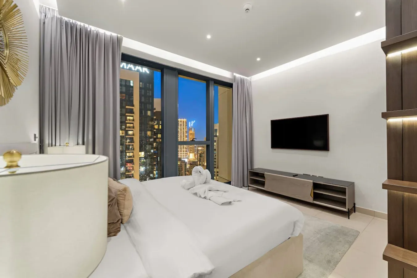 A bright bedroom with a bed, television, and floor-to-ceiling windows overlooking city lights.