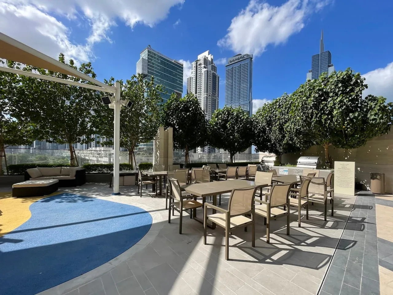 An outdoor patio with tables and seating beneath trees, with city towers in the background.