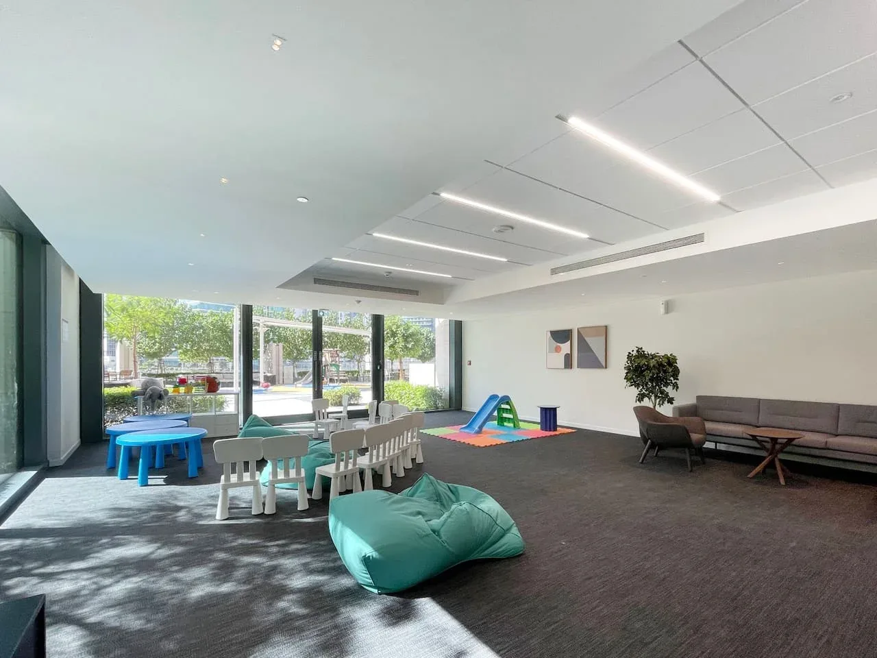 A bright indoor lounge has low seating, a children’s play area, and large windows.