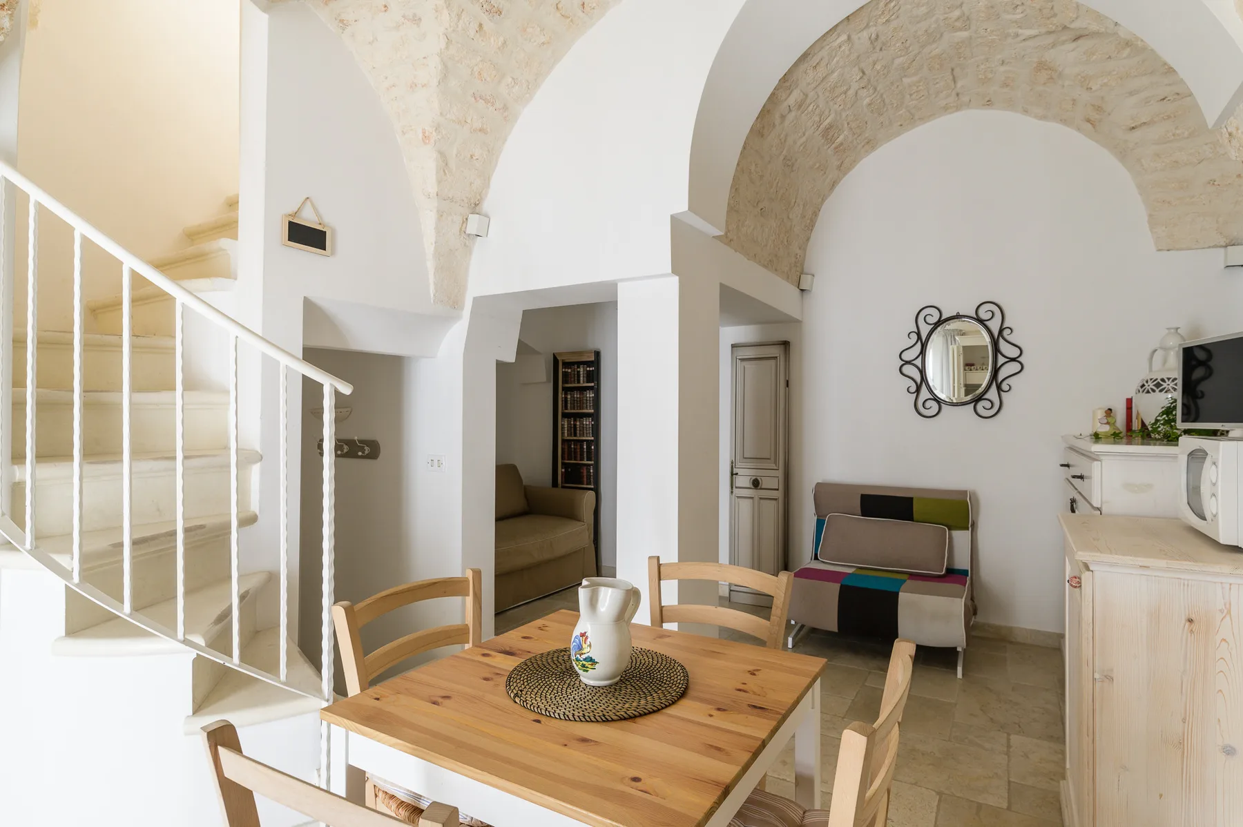 A dining area with a wooden table sits beside a small kitchen and staircase.