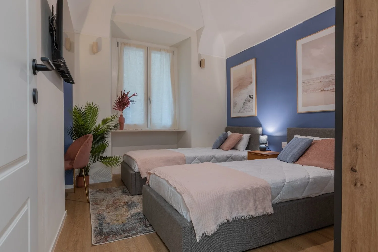A bedroom with two beds, a blue accent wall, and a window with sheer curtains.