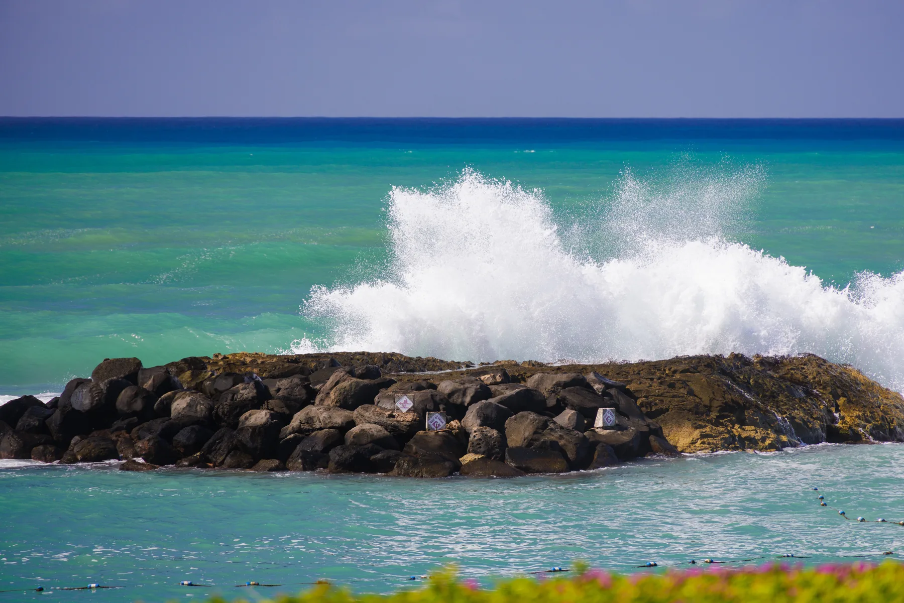 Las olas del mar rompen sobre un rompeolas rocoso junto al agua turquesa.