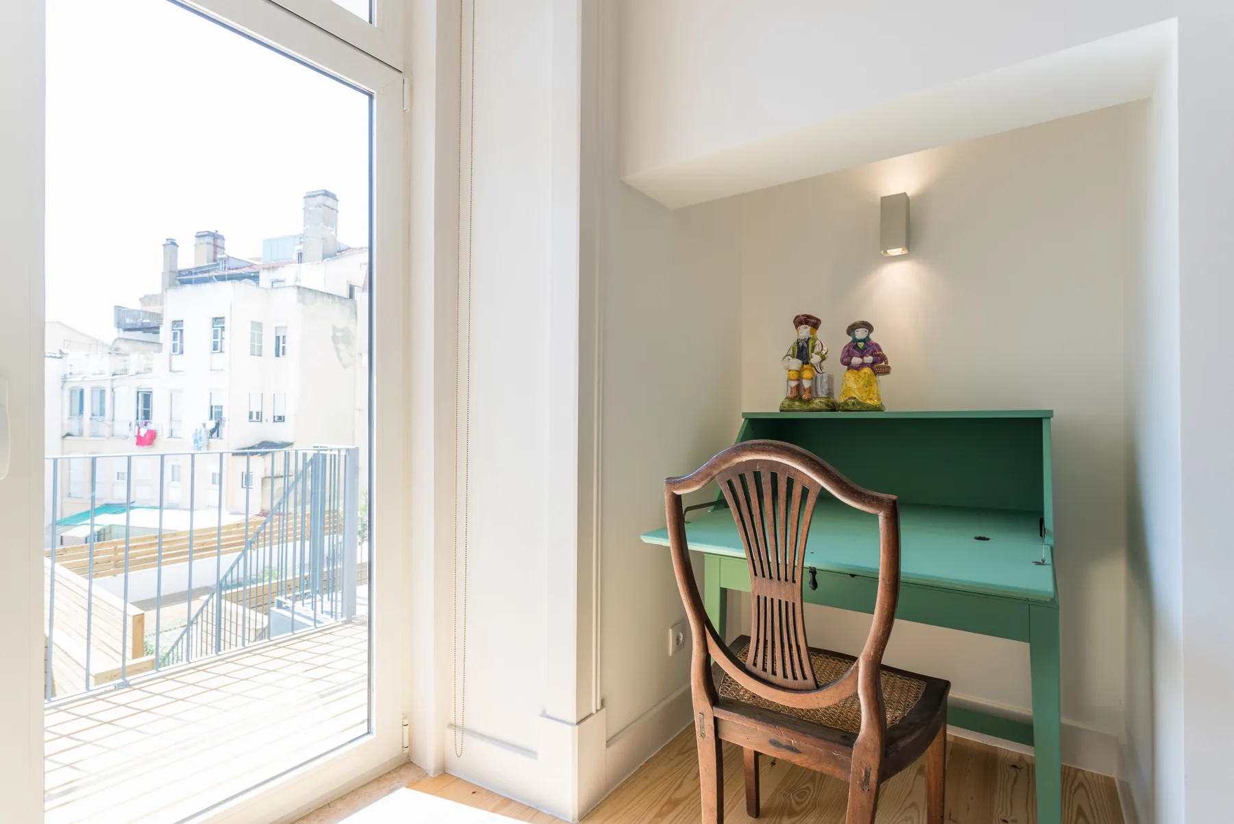 Un pequeño escritorio y un asiento de madera están junto a una ventana luminosa con vistas a edificios cercanos.