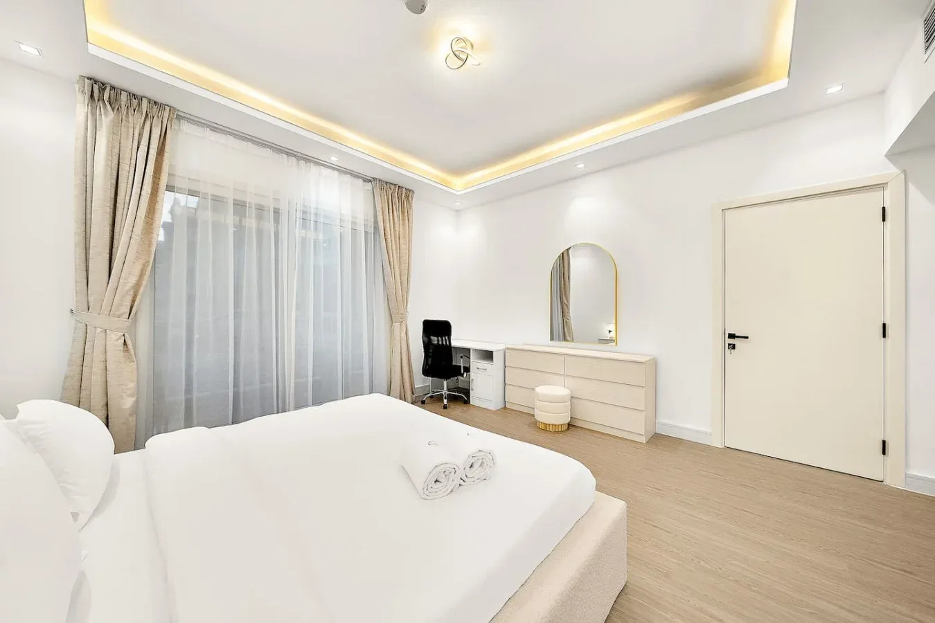 Un dormitorio luminoso con una cama arreglada, un escritorio y una gran ventana con cortinas.