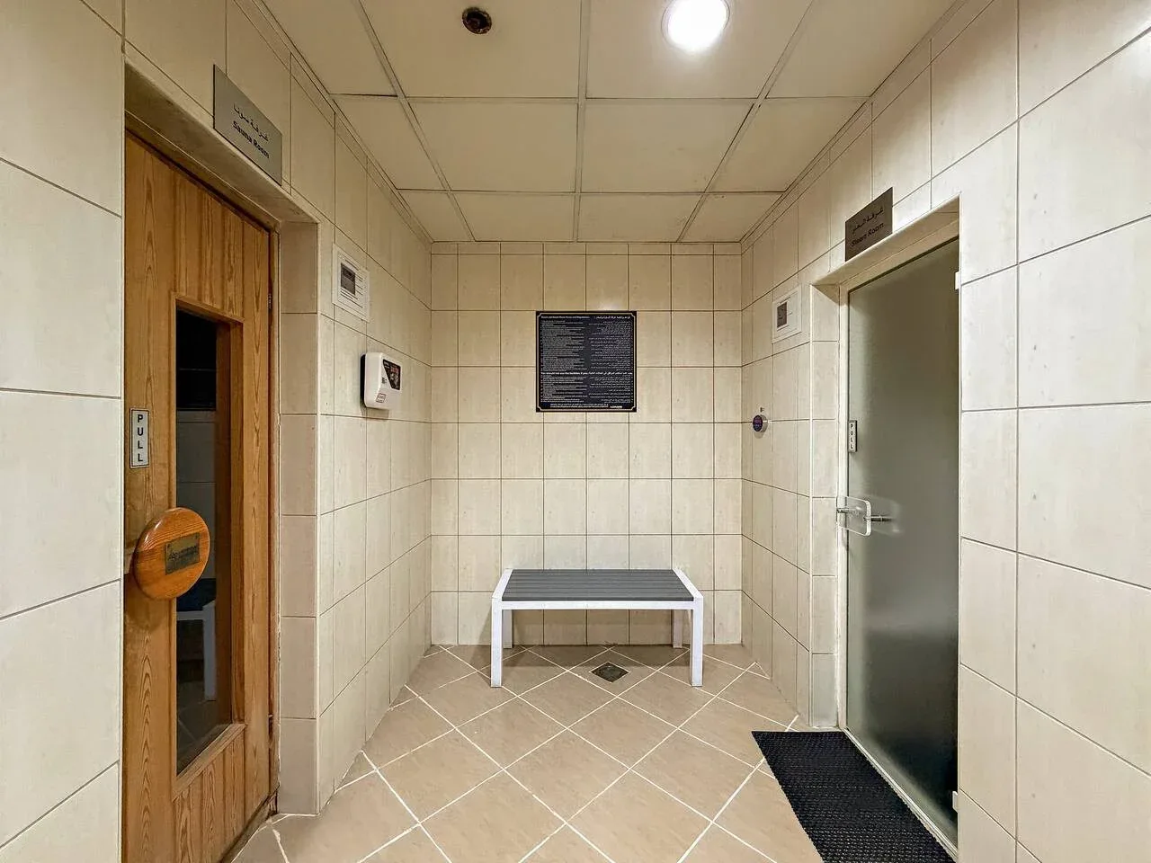 Pasillo interior con azulejos, una puerta de madera y un banco junto a la pared del fondo.