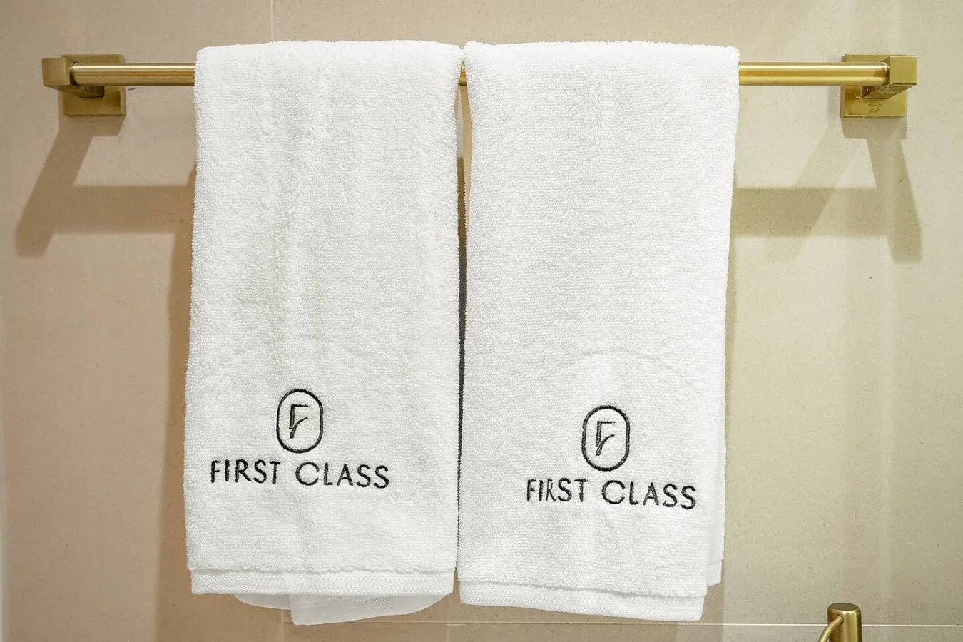Dos toallas blancas cuelgan de una barra dorada contra una pared beige de baño.