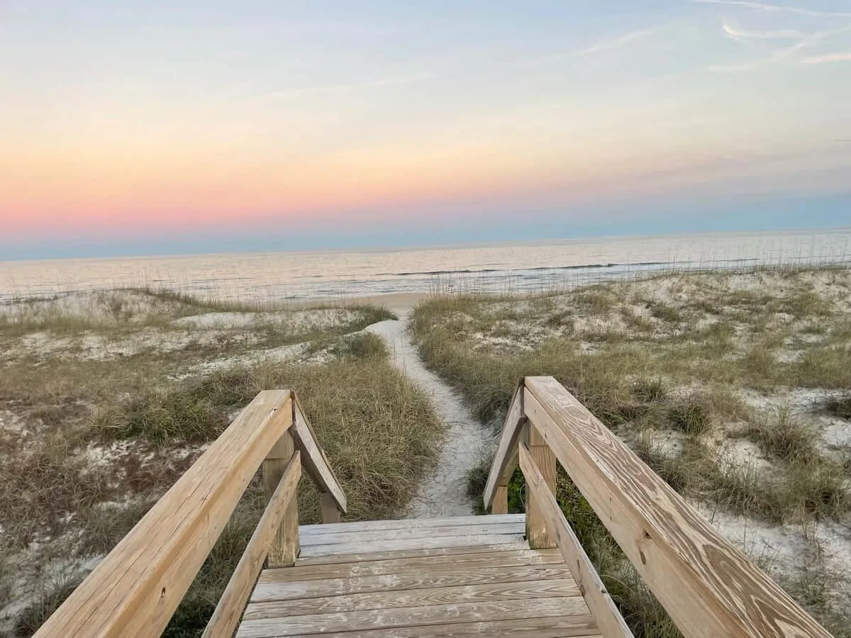 A wooden boardwalk crosses grassy dunes toward the ocean beneath a pastel sunset sky.