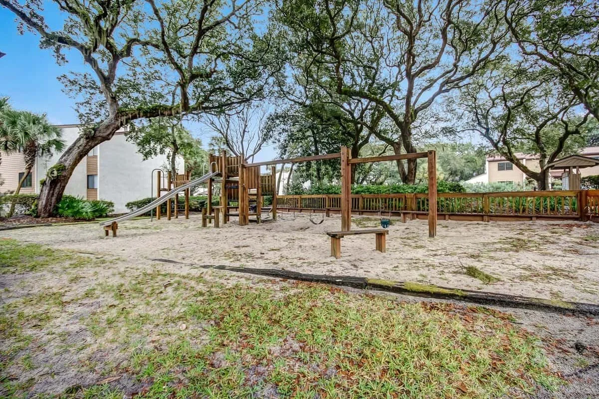 A playground with wooden climbing structures and swings beneath large trees.
