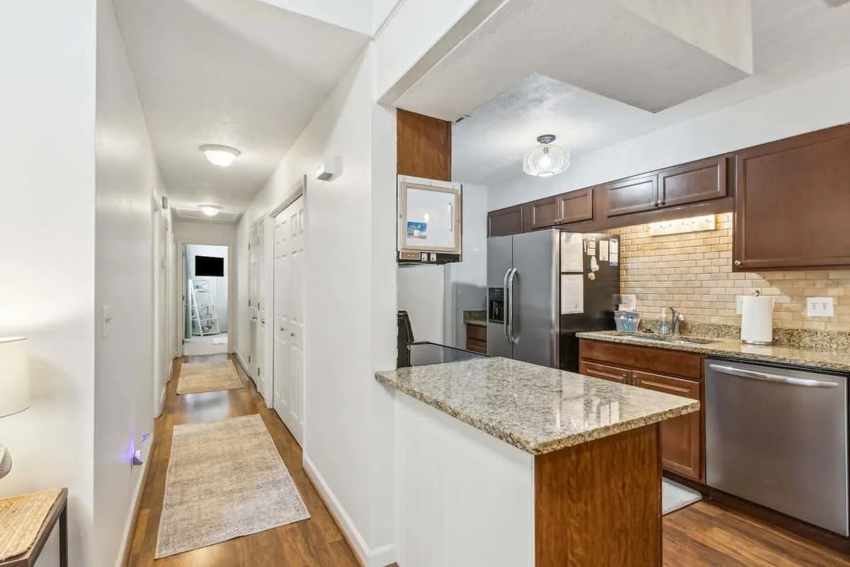 A kitchen with dark cabinets and a granite island opens onto a narrow hallway.
