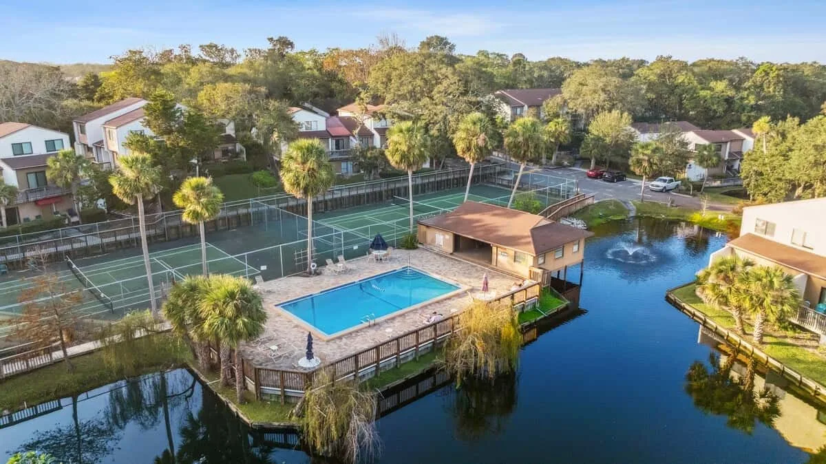 Aerial view of a pool, tennis courts, and buildings beside a pond.