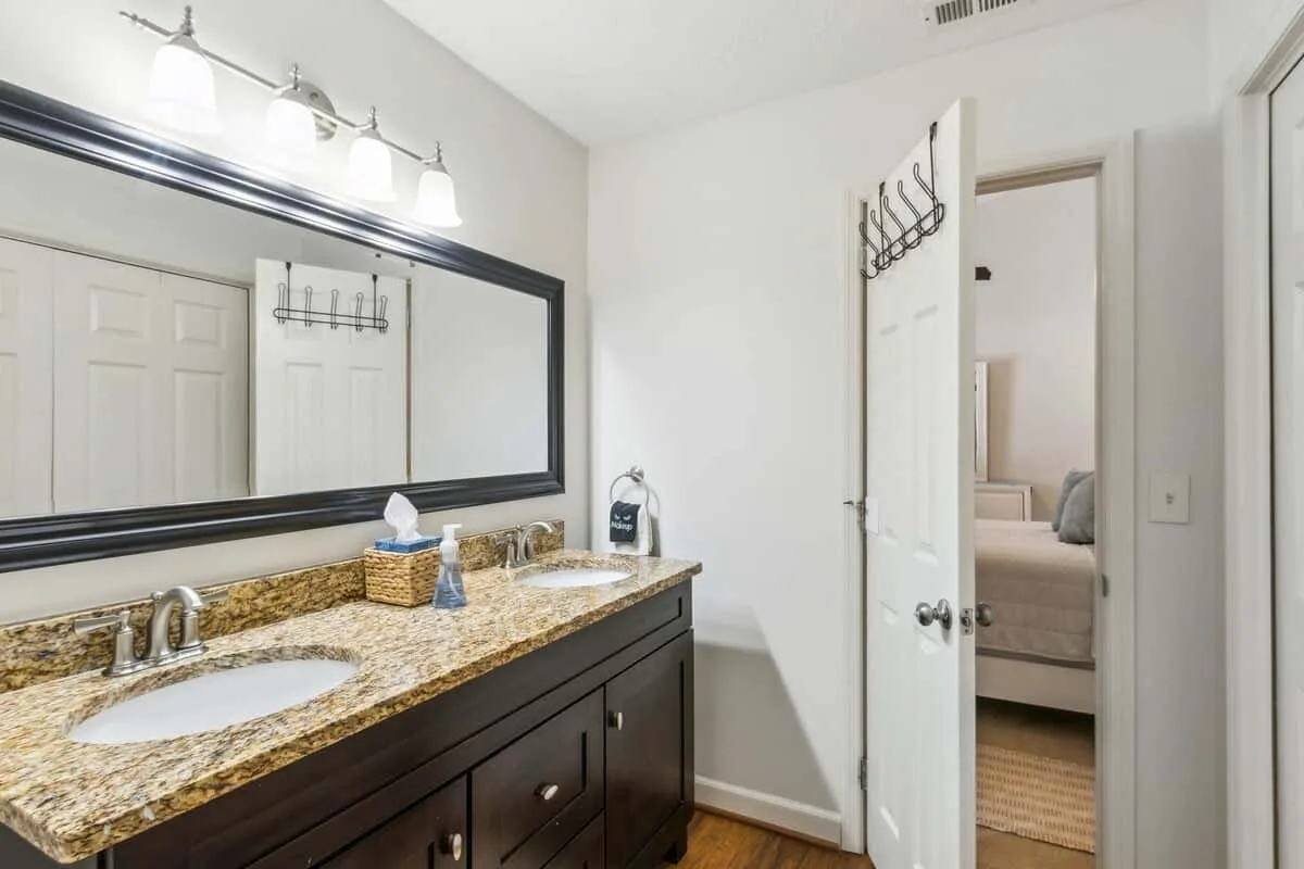 A bathroom vanity with a broad mirror and an open door to a bedroom.