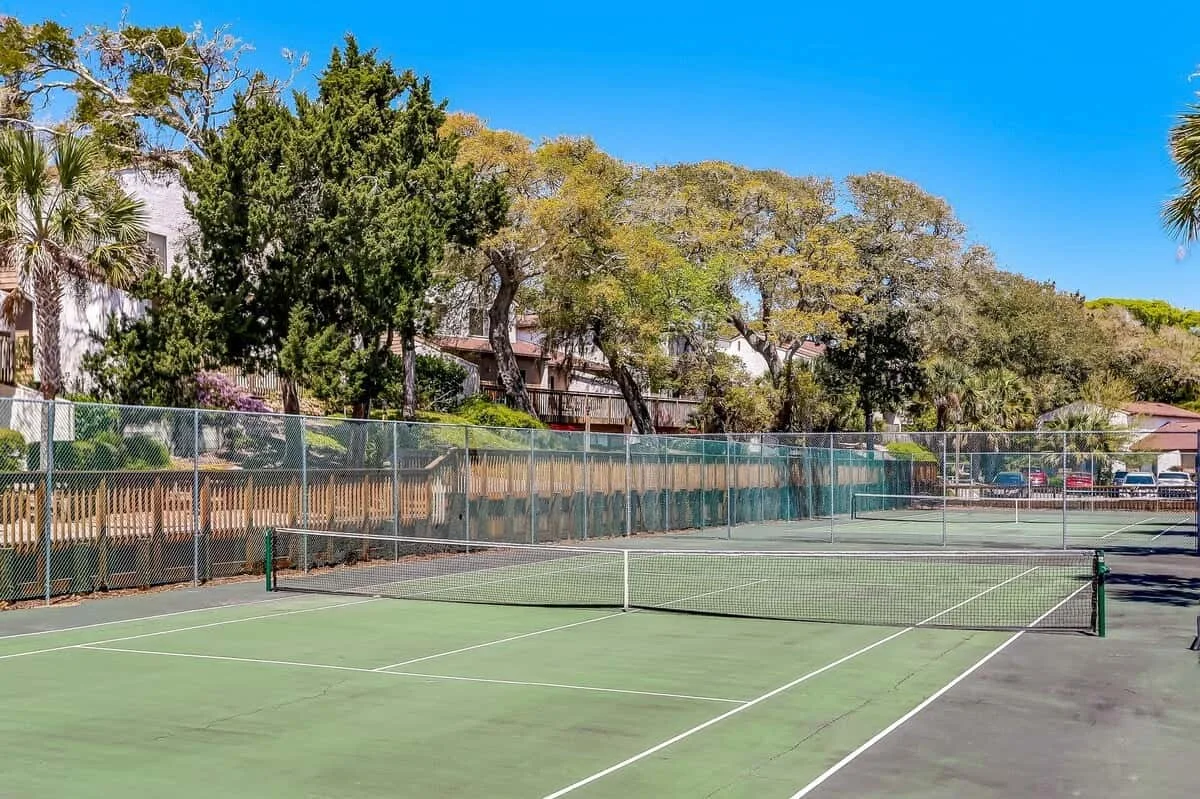 Outdoor tennis court with a net, surrounded by trees and fencing.