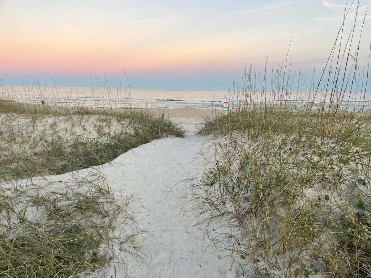 A sandy path through beach grass leads toward the shoreline at sunset.