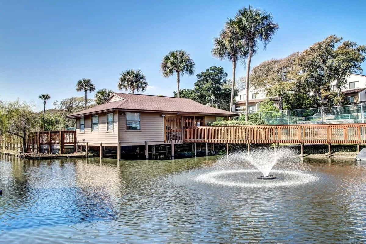 A waterside building stands beside a pond with a fountain and a wooden walkway.